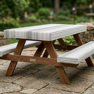 May include: A wooden picnic table with a brown finish and a small metal plaque labeled PicnicCo. The table features thick, rectangular cushions on the tabletop and both attached benches, all covered in a grey and white vertical striped fabric.