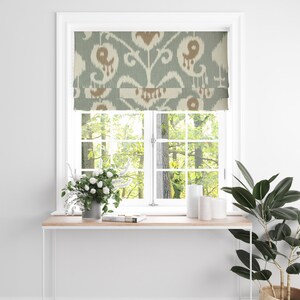 May include: A window with a roman blind featuring a blue and brown ikat pattern. The blind is pulled up, revealing a view of trees outside the window. There are white candles and a vase of white flowers on a white table below the window.