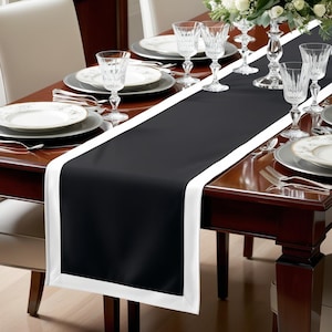 May include: Black and white table runner with a white border. The runner is on a wooden table with a white tablecloth and silver silverware.