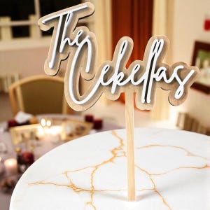 A wooden cake topper with the name "Thecefas" written on it, placed on a marble table.
