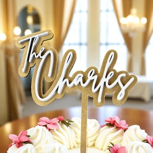 A gold-colored cake topper with the name "The Chairs" written on it, placed on top of a white cake with pink flowers.