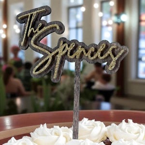 A wooden cake topper with the name "The Spence" written on it, placed on top of a white frosted cake.