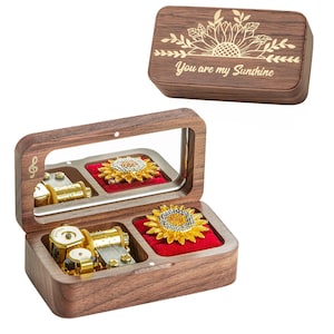 Puede incluir: Una caja de música de madera con interior espejado, con un mecanismo musical dorado y un soporte para joyas en forma de girasol. La tapa de la caja está grabada con un diseño de girasol y el texto "You are my Sunshine."