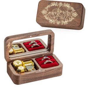 May include: A wooden music box with a hinged lid, featuring a mirrored interior and compartments for rings. The box is brown with a gold-colored musical mechanism and red velvet ring holders. The lid has an engraved design.