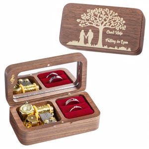 May include: A wooden music box with a mirrored interior, holding two rings in a red velvet compartment. The box features a gold-colored music mechanism and a treble clef. The lid has a tree design with the text "Can't Help Falling in Love".