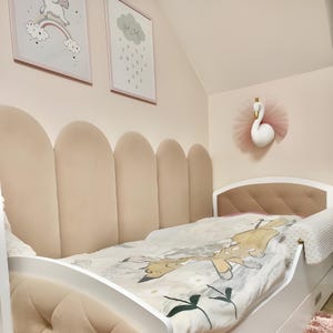 Könnte beinhalten: Ein weißes, gepolstertes Bett mit einem hellrosa Kopfteil mit geschwungenem Design. Das Bett hat einen weißen und grauen, gemusterten Bettbezug und ein weißes Kissen. An der Wand hinter dem Bett hängen zwei gerahmte Drucke, einer mit einem Einhorn und einem Regenbogen und der andere mit einer Wolke und Regentropfen. Ein weißer Schwan mit einem rosa Tüllrock hängt an der Wand.