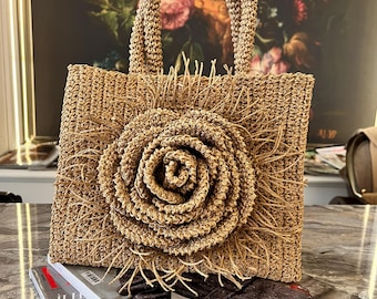 May include: A woven straw tote bag with a large, spiral-shaped flower detail. The bag has a rectangular shape with a woven handle and fringe accents. The bag is sitting on top of a magazine.