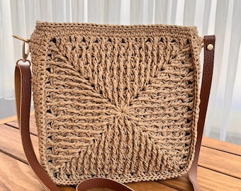 May include: A woven, square-shaped handbag in a light brown hue. The bag features a detailed, open weave pattern and a brown leather shoulder strap. The strap is adjustable and attaches to the bag with gold-colored hardware. The bag is sitting on a wooden surface.