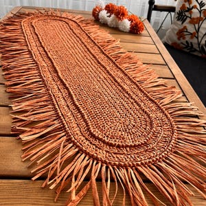 Peut inclure: Chemin de table ovale tissé, de couleur orange chaleureux. Le chemin de table présente une bordure à franges et une surface texturée, ajoutant une touche rustique. Il est posé sur une table en bois, mettant en valeur son design et sa texture.
