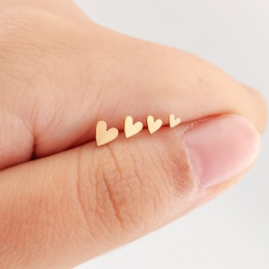 May include: A set of four small gold-colored heart-shaped studs arranged in a row by size from largest to smallest. Each heart features a smooth, flat surface and a simple, clean outline, presented as a collection of individual jewelry pieces.