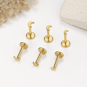 May include: Six gold-colored lip or ear studs. Three studs have a crescent moon design, and the other three have a flat, round top. The studs are arranged on a white surface with a neutral background.