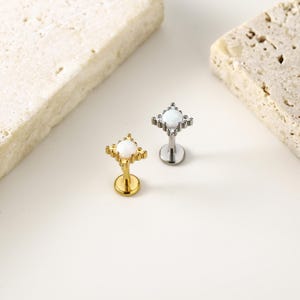 May include: Two lip piercing studs. One is gold-toned, the other silver-toned. Each features a white, round gemstone set in a square frame with decorative accents. The studs are displayed on a white surface.