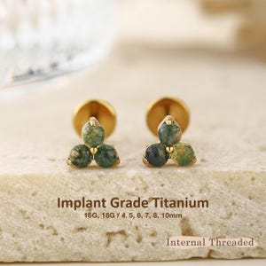 May include: Two gold-tone stud earrings featuring a cluster of three round moss agate stones. The text reads Implant Grade Titanium, 16G, 18G / 4, 5, 6, 7, 8, 10mm, and Internal Threaded. The earrings have a flat back post design.