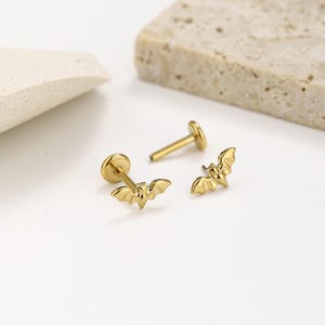 May include: Two gold-colored stud earrings featuring a small bat-shaped design. Each earring consists of a flat circular backing attached to a straight post, with the bat motif positioned at the front. The metal has a smooth, polished finish.