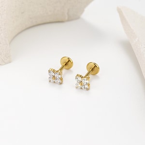 May include: Two gold-colored stud earrings. Each earring features a small, four-petal flower design, with clear, sparkling stones set in the center of each petal. The earrings have a flat, round backing. The background is white.