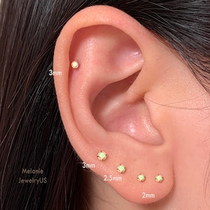 May include: Close-up of an ear with multiple small, round, opal-like stud earrings. The earrings are set in gold and vary in size, with measurements of 2mm, 2.5mm, and 3mm. The ear is against a neutral background.