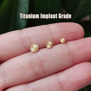 May include: Three small, gold-colored, round earrings with a sunburst design are displayed. The earrings are of varying sizes and are held in a hand. The text "Titanium Implant Grade" is above the earrings.