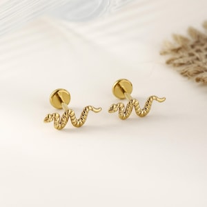 May include: A pair of gold-colored snake-shaped earrings. Each earring features a textured snake design with a flat, round backing. The earrings are displayed on a white surface, showcasing their intricate details and shiny finish.