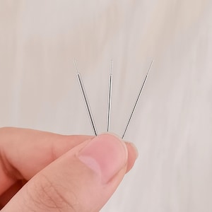 May include: Three slender, silver-toned metal acupuncture needles arranged in a fan shape. Each needle features a thin, straight shaft with a slightly thicker handle section at the base, designed for precision and professional use in therapeutic practice.