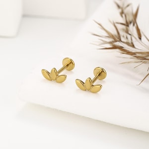 May include: Two gold-colored, flat-back earrings. Each earring features a three-petal design. The earrings have a post and a flat disc backing. The earrings are displayed on a white surface.