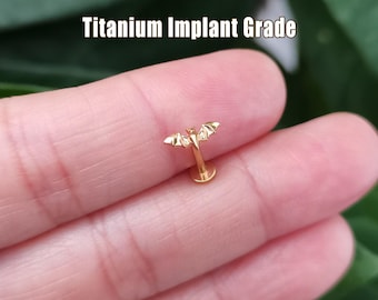 May include: A gold-colored bat-shaped piercing stud. The text "Titanium Implant Grade" is displayed above the stud. The bat has outstretched wings and is held between fingers.