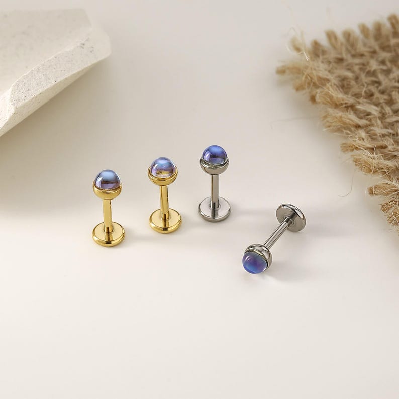 以下が含まれることがあります： 小さな虹色の青紫色のオーブが上部にある4つのジュエリー。3つはポストとベースがあり、2つは金色、1つは銀色です。4つ目のジュエリーは横に倒れています。