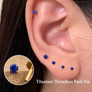 May include: Ear with multiple blue gemstone stud earrings. The earrings are in various sizes and are set in gold-colored metal. The text "Titanium Threadless Push Pin" is at the bottom of the image. A close-up of a single earring is in the lower left corner.