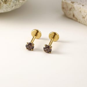 May include: Two gold-colored stud earrings with a small, round, dark purple gemstone. The earrings have a flat disc backing and a slender post. The gemstones are set in a four-prong setting.