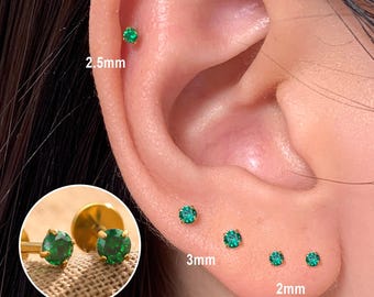 May include: A close-up of an ear with multiple small, round, emerald-green stud earrings. The earrings are set in gold-colored metal. Sizes are 2mm, 2.5mm, and 3mm. The earrings are displayed on an ear.