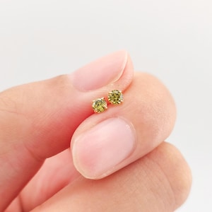 May include: A pair of small, round stud earrings with a light green gemstone set in a gold metal. The earrings are held in a person's hand against a white background. The gemstones have a slight sparkle.