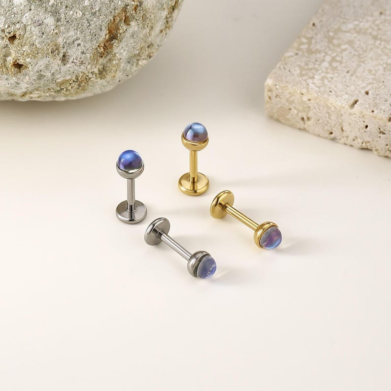 以下が含まれることがあります： 4つのジュエリー。銀色のものが2つ、金色のものが2つ。それぞれに小さく丸い青紫色の石が付いています。ジュエリーはボディピアス用に設計されており、裏面は平らでポストが付いています。白い表面に配置されています。