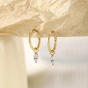 May include: A pair of gold-colored hoop earrings with a dangling, teardrop-shaped, clear gemstone. The earrings are displayed against a neutral, textured background, highlighting the jewelry's design and sparkle. The earrings are a classic and elegant accessory.