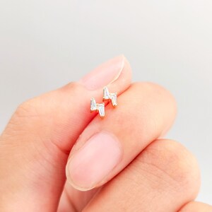 May include: A pair of gold-colored stud earrings in a lightning bolt design. Each earring features a series of small, clear, rectangular-cut stones. The earrings are held in a person's hand against a soft, neutral background.