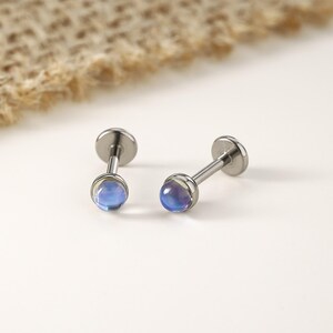 以下が含まれることがあります： イリデッセントブルーパープルの宝石が付いた、2つのシルバーカラーのリップピアススタッド。スタッドは片側に平らなディスク、もう一方に丸い宝石が付いています。宝石は微妙な色のグラデーションを持っています。