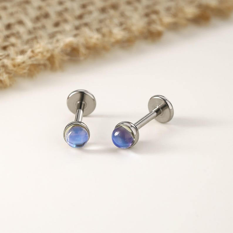 以下が含まれることがあります： イリデッセントブルーパープルの宝石が付いた、2つのシルバーカラーのリップピアススタッド。スタッドは片側に平らなディスク、もう一方に丸い宝石が付いています。宝石は微妙な色のグラデーションを持っています。