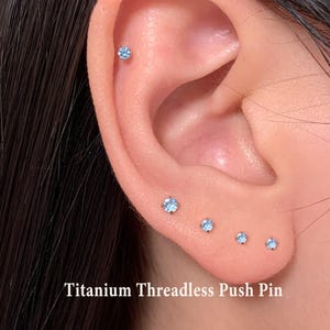 May include: Close-up of an ear with multiple small, light blue titanium threadless push pin earrings. The text "Titanium Threadless Push Pin" is visible at the bottom of the image. The earrings are arranged in a line and are of varying sizes.
