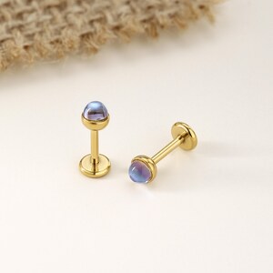 以下が含まれることがあります： イリデッセントな青紫色のカボションストーンが付いた、2つの金色の唇または軟骨イヤリング。イヤリングは平らなディスクベースと丸みを帯びたトップのポストを備えています。背景はニュートラルホワイトです。
