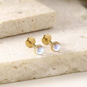 以下が含まれることがあります： イリデッセント、丸い、水色の宝石が付いた2つのゴールドスタッドピアス。イヤリングは、質感のある明るい色の表面に表示されています。イヤリングはシンプルでエレガントなデザインです。