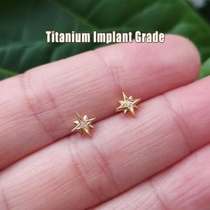 May include: A pair of gold-colored star-shaped earrings with a small, clear gem in the center of each. The earrings are labeled "Titanium Implant Grade".