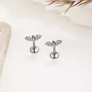 May include: A pair of silver-toned metal labret studs featuring a bat-shaped top design. Each piece consists of a flat circular base, a straight post, and a detailed bat silhouette with outstretched wings, crafted from a polished metallic material.