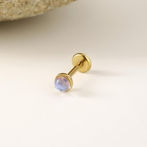 May include: A gold-colored, flat-backed earring with a light blue, round gemstone. The earring has a post and a flat disc on the back. The gemstone is set in a gold bezel.