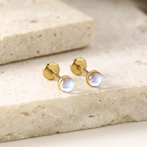 May include: A pair of gold-colored stud earrings with iridescent, round, light blue gemstones. The earrings are displayed on a textured, light beige surface, showcasing their delicate design and subtle color variations. The earrings are a type of jewelry.