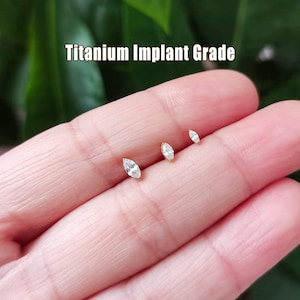May include: Three gold-toned stud earrings featuring marquise-cut clear stones of varying sizes, arranged in a row. The text Titanium Implant Grade is displayed at the top of the image. Each earring has a metallic post and a clear faceted stone.