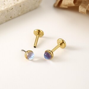 以下が含まれることがあります： イリデッセントな丸い宝石が付いた、3つのゴールドカラーのリップピアススタッド。2つのスタッドは上部に平らなディスクがあり、1つは湾曲したポストが付いています。宝石は青、紫、透明色の混合色です。ジュエリーは白い表面にあります。