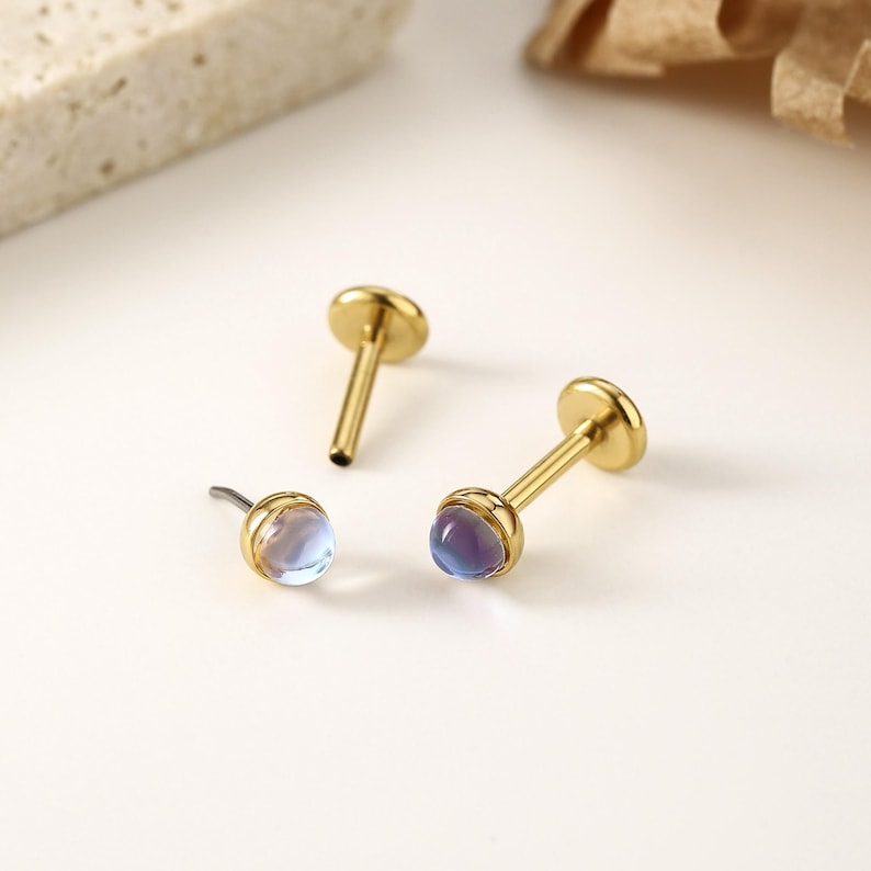 以下が含まれることがあります： イリデッセントな丸い宝石が付いた、3つのゴールドカラーのリップピアススタッド。2つのスタッドは上部に平らなディスクがあり、1つは湾曲したポストが付いています。宝石は青、紫、透明色の混合色です。ジュエリーは白い表面にあります。