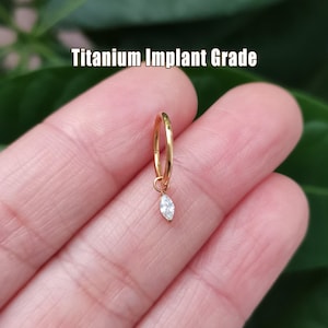 May include: A gold-colored hoop earring with a dangling, marquise-cut, clear gemstone. The text "Titanium Implant Grade" is displayed above the earring. The earring is held in a hand against a blurred green background.
