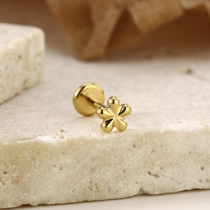 May include: A gold-colored flower-shaped earring stud. The earring has a flat back and a post for piercing. The flower design has five petals. The earring is displayed on a textured, light-colored surface.