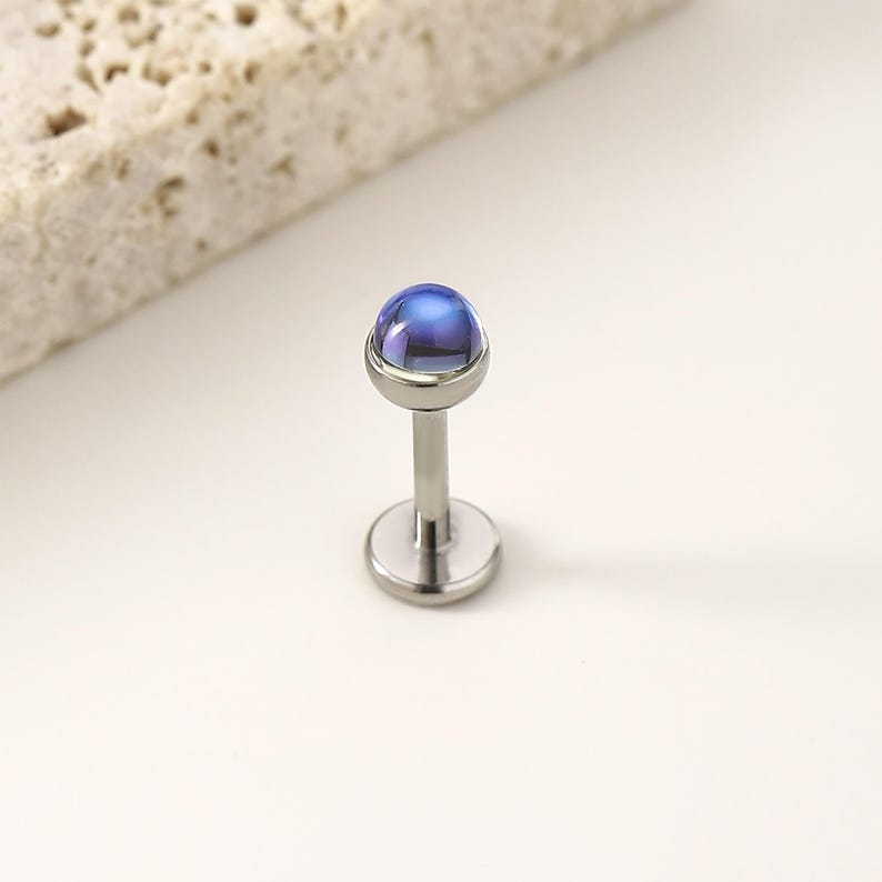 以下が含まれることがあります： 丸い青と紫の宝石が付いた、単一の銀色のリップピアススタッド。スタッドは平らなベースと長く細いポストを備えています。宝石はドーム型で光を反射し、きらめく効果を生み出します。ジュエリーはニュートラルな背景にセットされています。