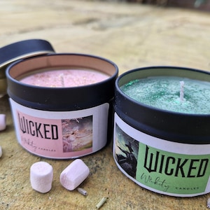 May include: Two round black tin candles with the word "WICKED" on the labels. One candle has pink wax with glitter, the other has green wax. Marshmallows are scattered around the candles. The candle lids are black.