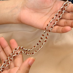 May include: A necklace featuring a series of small, round, reddish-brown rudraksha beads, each individually encased in a polished silver-toned metal setting, linked together to form a continuous chain of circular segments.